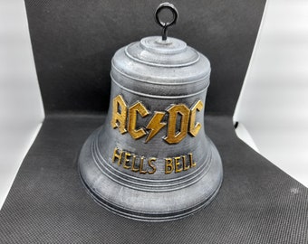 May include: A gray bell-shaped ornament with the text "AC/DC" in gold lettering and a lightning bolt. Below the band's name is the text "HELLS BELL". The bell has a loop at the top for hanging.