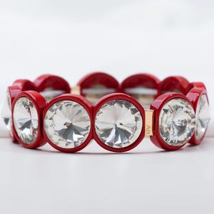 May include: A red bracelet with large, clear, round crystals. The bracelet features a series of red, circular links that encircle the crystals. The crystals are faceted, reflecting light. The bracelet is a statement piece of jewelry.