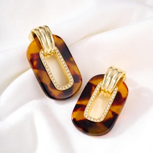 May include: A pair of tortoise shell-patterned earrings. Each earring features a gold-colored top and a rectangular shape with a gold-colored, crystal-studded center. The earrings are set against a white background.
