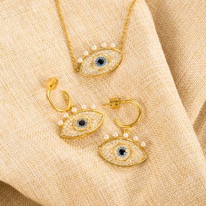 May include: A gold necklace, earrings, and bracelet set with a blue evil eye charm surrounded by white pearls. The jewelry is on a beige fabric background.