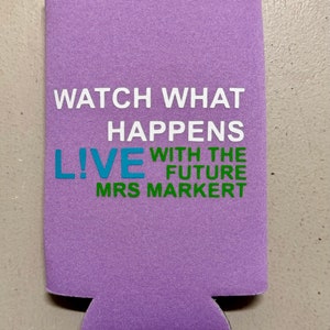 May include: A lavender-colored can cooler with the text "WATCH WHAT HAPPENS LIVE WITH THE FUTURE MRS MARKERT" in white, teal, and green. The cooler is designed to keep beverages cold and features a cut-out for easy gripping.