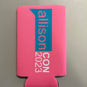 May include: Pink can cooler with the text "allison CON 2023" in white and teal. The cooler is made of a neoprene-like material and has a black bottom. The text is vertically aligned.