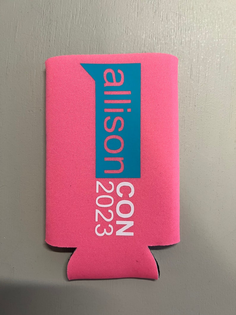 May include: Pink can cooler with the text "allison CON 2023" in white and teal. The cooler is made of a neoprene-like material and has a black bottom. The text is vertically aligned.