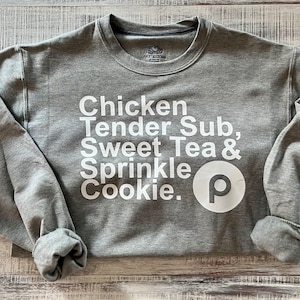 Puede incluir: Sudadera gris con texto blanco que dice "Chicken Tender Sub, Sweet Tea & Sprinkle Cookie." Un círculo blanco con una "P" dentro está al lado del texto.