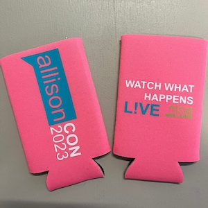 May include: Two pink can coolers with text. One cooler has "allison CON 2023" in white and teal. The other cooler has "WATCH WHAT HAPPENS LIVE" in white and teal, with a quote in green.