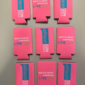 May include: Pink can coolers with teal and white text. The text includes "allison CON 2023" and "WATCH WHAT HAPPENS LIVE". The coolers are made of a foam material and are designed to keep beverages cold.