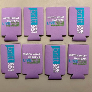 May include: Eight lavender can coolers with the text "britt CON 2024" and "WATCH WHAT HAPPENS LIVE WITH THE FUTURE MRS MARKERT" printed in white and teal. The coolers are made of a foam material.