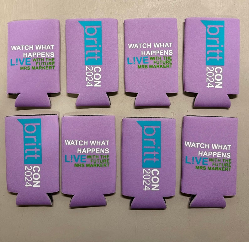 May include: Eight lavender can coolers with the text "britt CON 2024" and "WATCH WHAT HAPPENS LIVE WITH THE FUTURE MRS MARKERT" printed in white and teal. The coolers are made of a foam material.