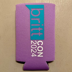 May include: A purple beverage can cooler with the text "britt CON 2024" in white and a teal blue accent. The cooler is made of a foam-like material and has a cutout at the bottom.