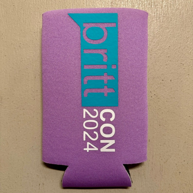 May include: A purple beverage can cooler with the text "britt CON 2024" in white and a teal blue accent. The cooler is made of a foam-like material and has a cutout at the bottom.
