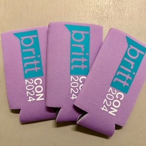 May include: Three lavender-colored can coolers with the text "britt CON 2024" printed in white and teal. The coolers are made of a foam-like material and are designed to keep beverages cold. The text is vertically aligned.