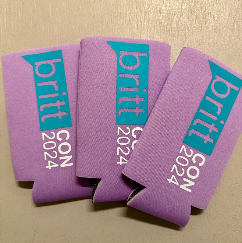 May include: Three lavender-colored can coolers with the text "britt CON 2024" printed in white and teal. The coolers are made of a foam-like material and are designed to keep beverages cold. The text is vertically aligned.