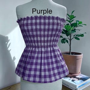 May include: A strapless tube top featuring a purple and white plaid pattern with a smocked, elasticized bodice and a ruffled top edge. The word Purple is printed in black text above the garment, which is displayed on a mannequin torso.