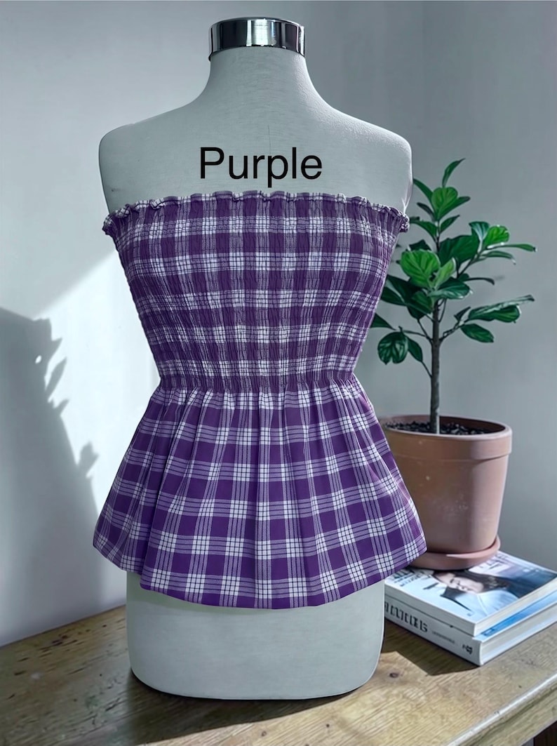 May include: A strapless tube top featuring a purple and white plaid pattern with a smocked, elasticized bodice and a ruffled top edge. The word Purple is printed in black text above the garment, which is displayed on a mannequin torso.