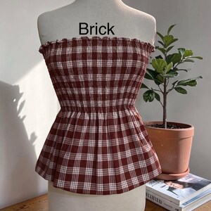 Peut inclure: Un haut bustier sans bretelles &agrave; motif &eacute;cossais rouge brique et blanc. Le v&ecirc;tement pr&eacute;sente un corsage smock&eacute; et &eacute;lastique avec un bord sup&eacute;rieur volant&eacute; et un ourlet &agrave; basque &eacute;vas&eacute;. Le mot Brick est imprim&eacute; en noir au-dessus du v&ecirc;tement.