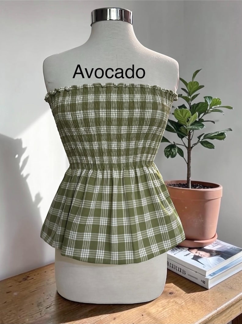 Peut inclure: Un haut bustier sans bretelles &agrave; motif &eacute;cossais vert olive et blanc. Le v&ecirc;tement pr&eacute;sente un corsage smock&eacute; et &eacute;lastique avec un bord sup&eacute;rieur volant&eacute; et un ourlet &agrave; basque &eacute;vas&eacute;. Le mot Avocado est imprim&eacute; en noir au-dessus du v&ecirc;tement.