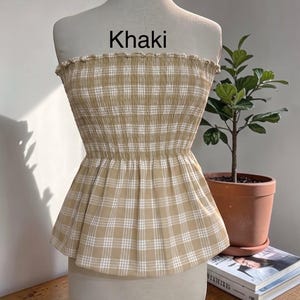 May include: A strapless tube top in a khaki and white plaid pattern. The garment features a smocked, elasticized bodice with a ruffled top edge and a flared peplum hem. The word Khaki is printed in black text above the garment on the mannequin.