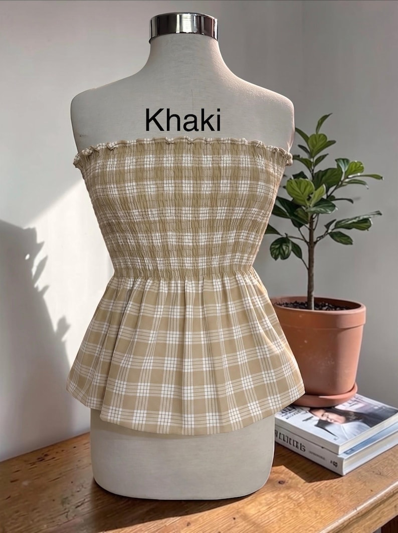 May include: A strapless tube top in a khaki and white plaid pattern. The garment features a smocked, elasticized bodice with a ruffled top edge and a flared peplum hem. The word Khaki is printed in black text above the garment on the mannequin.
