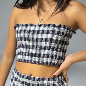 Könnte beinhalten: Ein blau-weiß kariertes Tube-Top mit gerüschtem Rand. Das Oberteil besteht aus einem dehnbaren Material und ist ohne Träger konzipiert. Das Oberteil ist Teil eines passenden Sets.