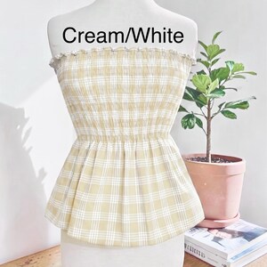 May include: A strapless tube top featuring a cream and white plaid pattern. The garment has a smocked, elasticized bodice that creates a gathered texture, transitioning into a flared peplum hem. The text "Cream/White" is printed on the chest area.