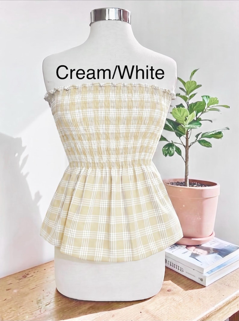 May include: A strapless tube top featuring a cream and white plaid pattern. The garment has a smocked, elasticized bodice that creates a gathered texture, transitioning into a flared peplum hem. The text "Cream/White" is printed on the chest area.