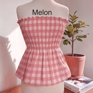 Peut inclure: Un haut bustier sans bretelles &agrave; motif &eacute;cossais rose poudr&eacute; et blanc. Le v&ecirc;tement pr&eacute;sente un corsage fronc&eacute; et &eacute;lastiqu&eacute; avec un bord sup&eacute;rieur volant&eacute; et un ourlet &agrave; basque &eacute;vas&eacute;. Le mot "Melon" est imprim&eacute; en noir au-dessus du haut.