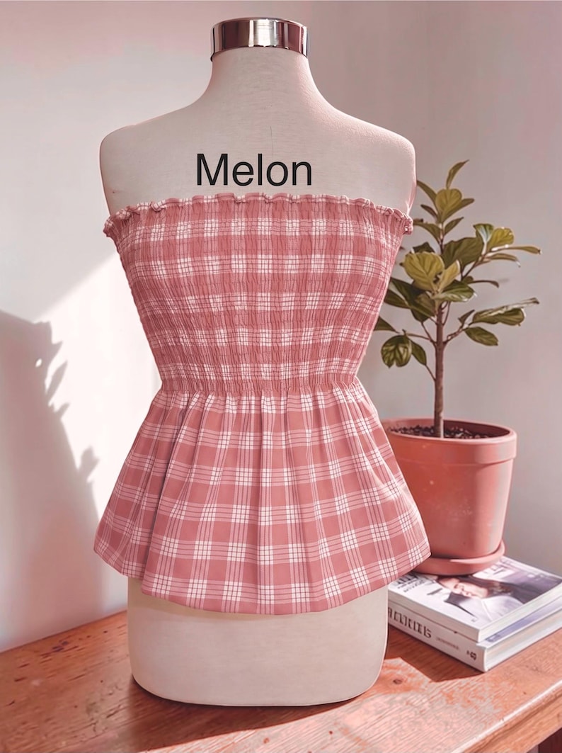Peut inclure: Un haut bustier sans bretelles &agrave; motif &eacute;cossais rose poudr&eacute; et blanc. Le v&ecirc;tement pr&eacute;sente un corsage fronc&eacute; et &eacute;lastiqu&eacute; avec un bord sup&eacute;rieur volant&eacute; et un ourlet &agrave; basque &eacute;vas&eacute;. Le mot "Melon" est imprim&eacute; en noir au-dessus du haut.