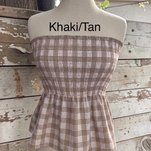 May include: A strapless tube top in a khaki and tan plaid pattern with a smocked bodice and a flared peplum hem. The garment features a white grid design over the brown fabric and includes the text Khaki/Tan printed above the neckline.