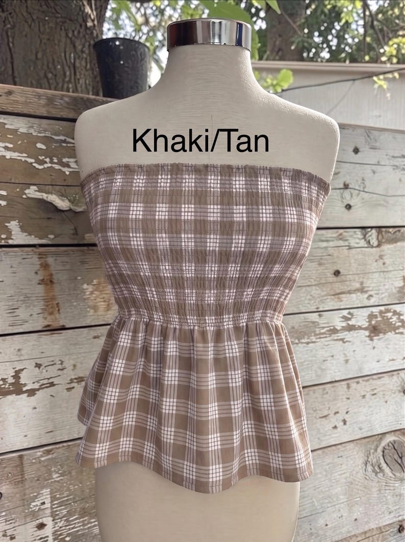 May include: A strapless tube top in a khaki and tan plaid pattern with a smocked bodice and a flared peplum hem. The garment features a white grid design over the brown fabric and includes the text Khaki/Tan printed above the neckline.