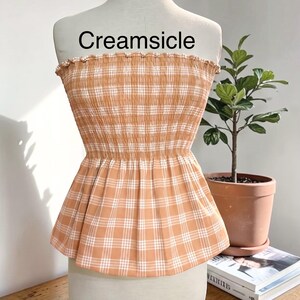 Peut inclure: Un haut &agrave; basque sans bretelles avec un motif &agrave; carreaux orange clair et blanc. Le corsage pr&eacute;sente un design smock&eacute; et &eacute;lastique avec un bord sup&eacute;rieur volant&eacute;. Le mot Creamsicle est imprim&eacute; en noir au-dessus du v&ecirc;tement sur le mannequin.