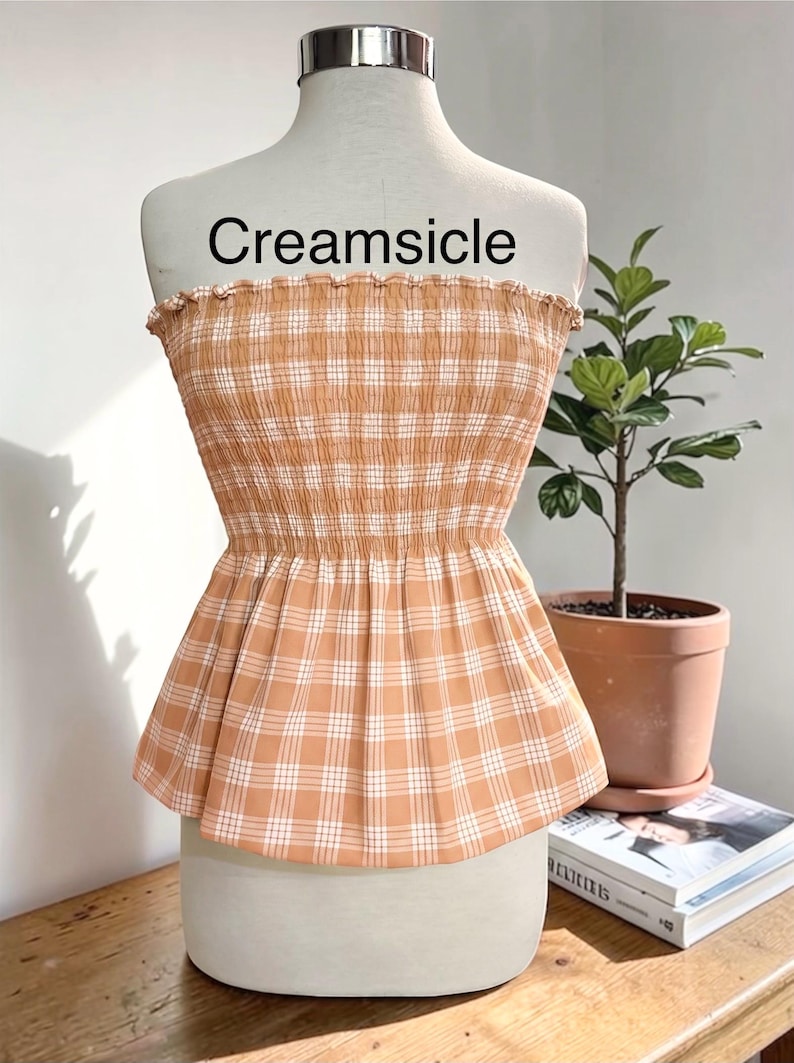 Peut inclure: Un haut &agrave; basque sans bretelles avec un motif &agrave; carreaux orange clair et blanc. Le corsage pr&eacute;sente un design smock&eacute; et &eacute;lastique avec un bord sup&eacute;rieur volant&eacute;. Le mot Creamsicle est imprim&eacute; en noir au-dessus du v&ecirc;tement sur le mannequin.
