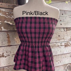 May include: A strapless tube top featuring a pink and black plaid pattern with a smocked elastic bodice and a flared peplum hem. The text Pink/Black is printed above the garment, which is displayed on a mannequin torso.