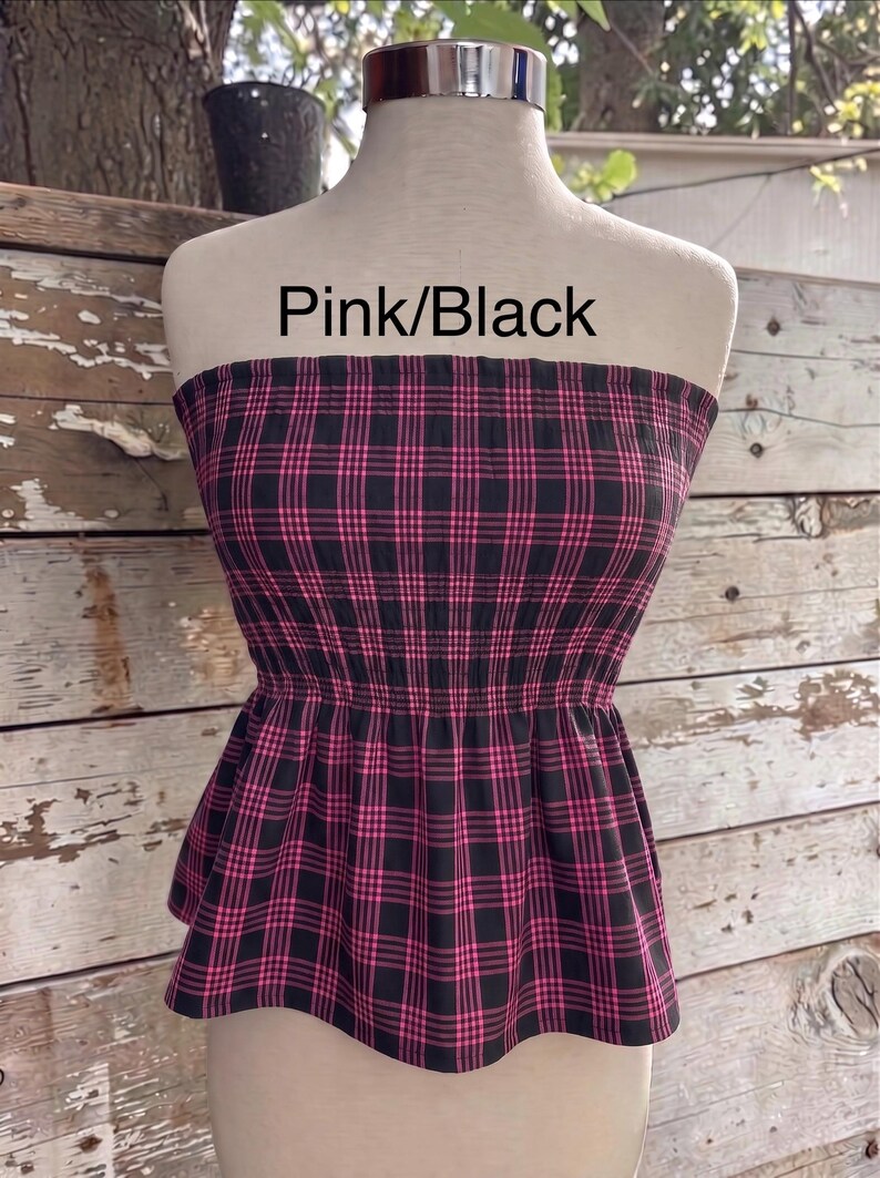 May include: A strapless tube top featuring a pink and black plaid pattern with a smocked elastic bodice and a flared peplum hem. The text Pink/Black is printed above the garment, which is displayed on a mannequin torso.