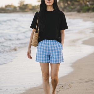 May include: A black short-sleeved t-shirt paired with blue and white checkered shorts featuring an elastic waistband and side pockets, accompanied by a woven straw tote bag with thin shoulder straps.