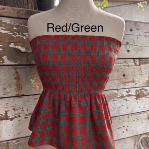 May include: A strapless tube top featuring a red and green plaid pattern with a smocked bodice and a flared peplum hem. The text Red/Green is printed above the garment on the mannequin. The fabric has a textured, crinkled appearance throughout.