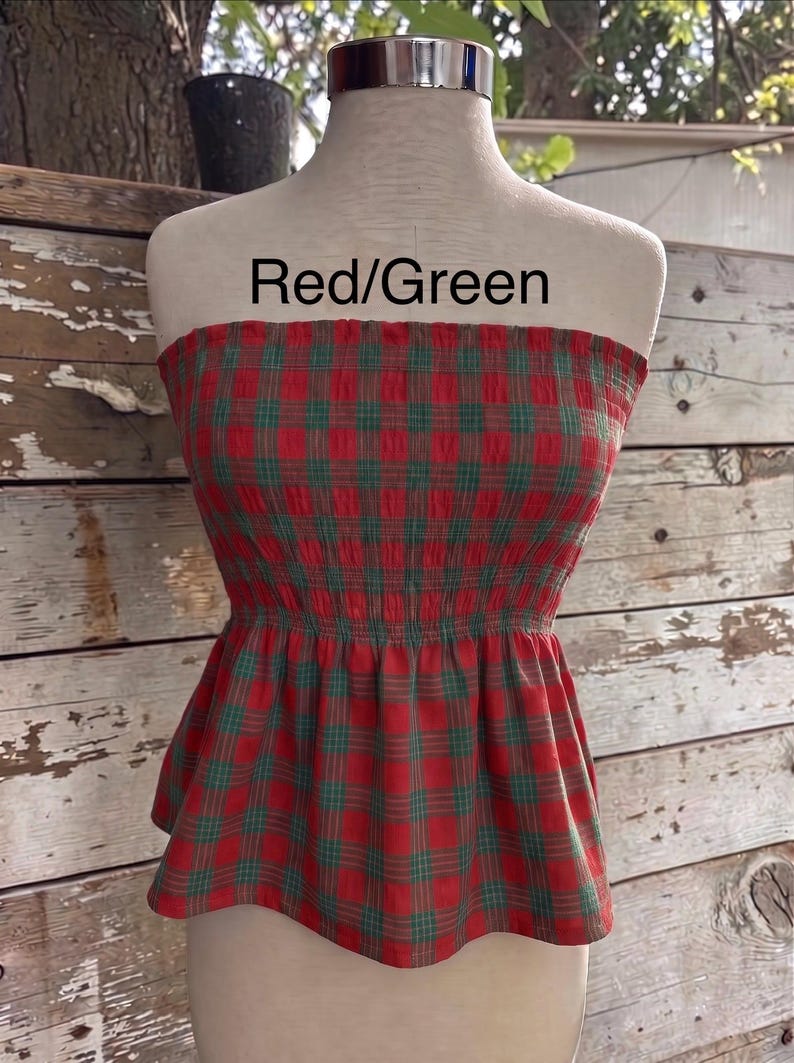 May include: A strapless tube top featuring a red and green plaid pattern with a smocked bodice and a flared peplum hem. The text Red/Green is printed above the garment on the mannequin. The fabric has a textured, crinkled appearance throughout.