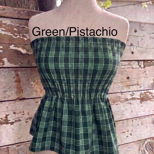 May include: A strapless tube top featuring a dark green and light green pistachio plaid pattern. The garment has a smocked, elasticized bodice that cinches at the waist and flares into a peplum hem. The text Green/Pistachio is visible.