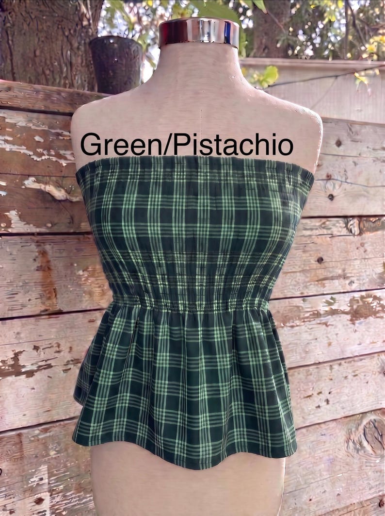May include: A strapless tube top featuring a dark green and light green pistachio plaid pattern. The garment has a smocked, elasticized bodice that cinches at the waist and flares into a peplum hem. The text Green/Pistachio is visible.