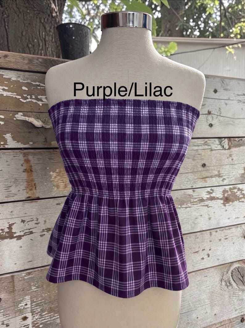 May include: A strapless tube top featuring a purple and white plaid pattern with a smocked bodice and a flared peplum hem. The text "Purple/Lilac" is printed above the garment on the mannequin.
