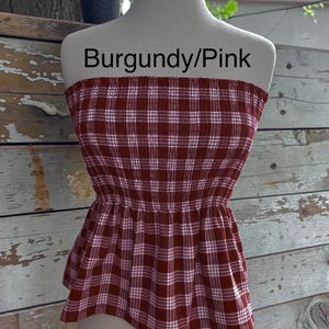 May include: A strapless tube top featuring a burgundy and pink plaid pattern with white grid lines. The garment has a smocked, elasticized bodice that transitions into a slightly flared peplum hem. The text Burgundy/Pink is visible.