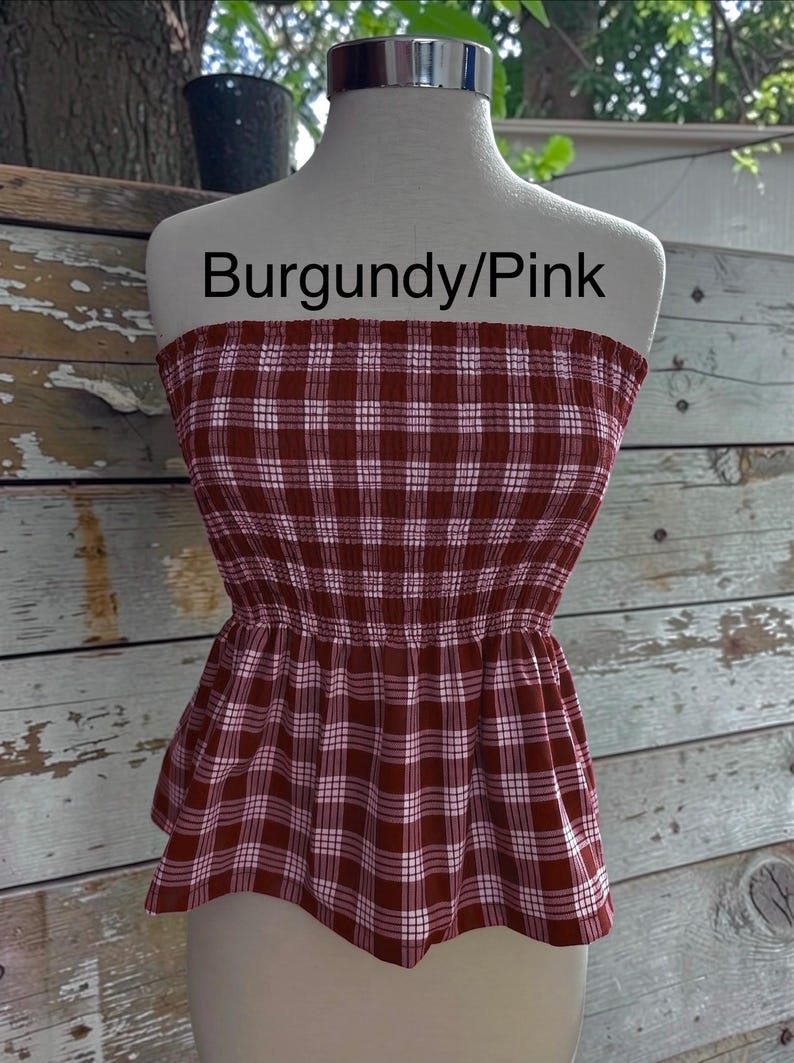 May include: A strapless tube top featuring a burgundy and pink plaid pattern with white grid lines. The garment has a smocked, elasticized bodice that transitions into a slightly flared peplum hem. The text Burgundy/Pink is visible.