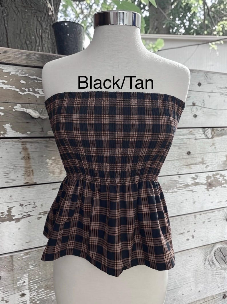 May include: A strapless tube top featuring a black and tan plaid pattern with a smocked bodice and a flared peplum hem. The text Black/Tan is printed above the garment on the mannequin.