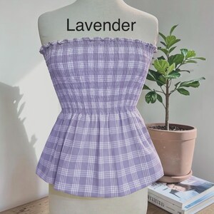 May include: A strapless tube top in a light lavender and white plaid pattern. The garment features a smocked, elasticized bodice with a ruffled top edge and a flared peplum hem. The word Lavender is printed in black text above the garment.