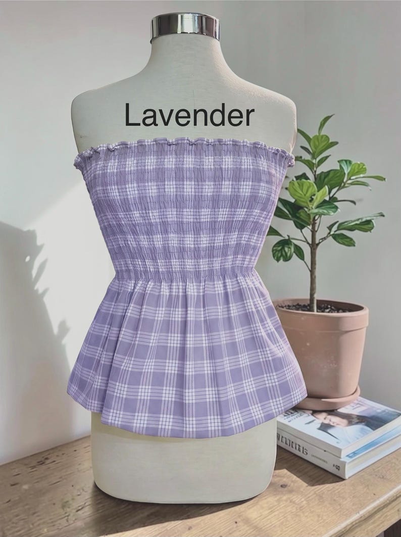 May include: A strapless tube top in a light lavender and white plaid pattern. The garment features a smocked, elasticized bodice with a ruffled top edge and a flared peplum hem. The word Lavender is printed in black text above the garment.