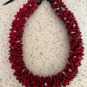 Könnte beinhalten: Ein leuchtend rotes Lei mit einem schwarzen Band. Das Lei besteht aus dicht gepackten, geschichteten roten Stoffblumen, die mit weißen und grünen Akzenten durchsetzt sind. Das Lei ist eine traditionelle hawaiianische Girlande, die oft zu Feiern getragen wird.