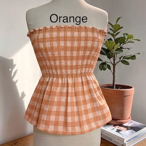 Peut inclure: Un haut bustier sans bretelles &agrave; motif &eacute;cossais orange clair et blanc. Le v&ecirc;tement pr&eacute;sente un corsage smock&eacute; et &eacute;lastique avec un bord sup&eacute;rieur volant&eacute; et un ourlet &agrave; basque &eacute;vas&eacute;. Le mot "Orange" est imprim&eacute; en noir au-dessus du haut.