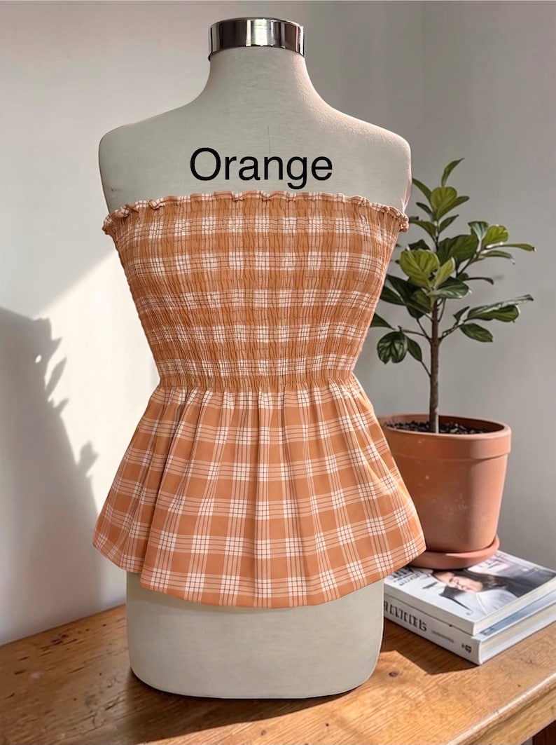 Peut inclure: Un haut bustier sans bretelles &agrave; motif &eacute;cossais orange clair et blanc. Le v&ecirc;tement pr&eacute;sente un corsage smock&eacute; et &eacute;lastique avec un bord sup&eacute;rieur volant&eacute; et un ourlet &agrave; basque &eacute;vas&eacute;. Le mot "Orange" est imprim&eacute; en noir au-dessus du haut.