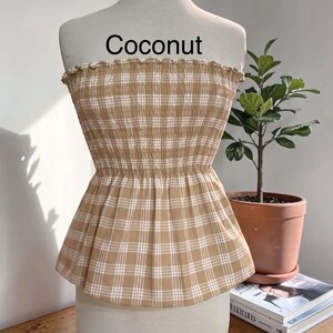 May include: A strapless tube top in a tan and white plaid pattern with a smocked, elasticized bodice and a ruffled top edge. The garment features the word Coconut printed in black text above the neckline on the display mannequin.