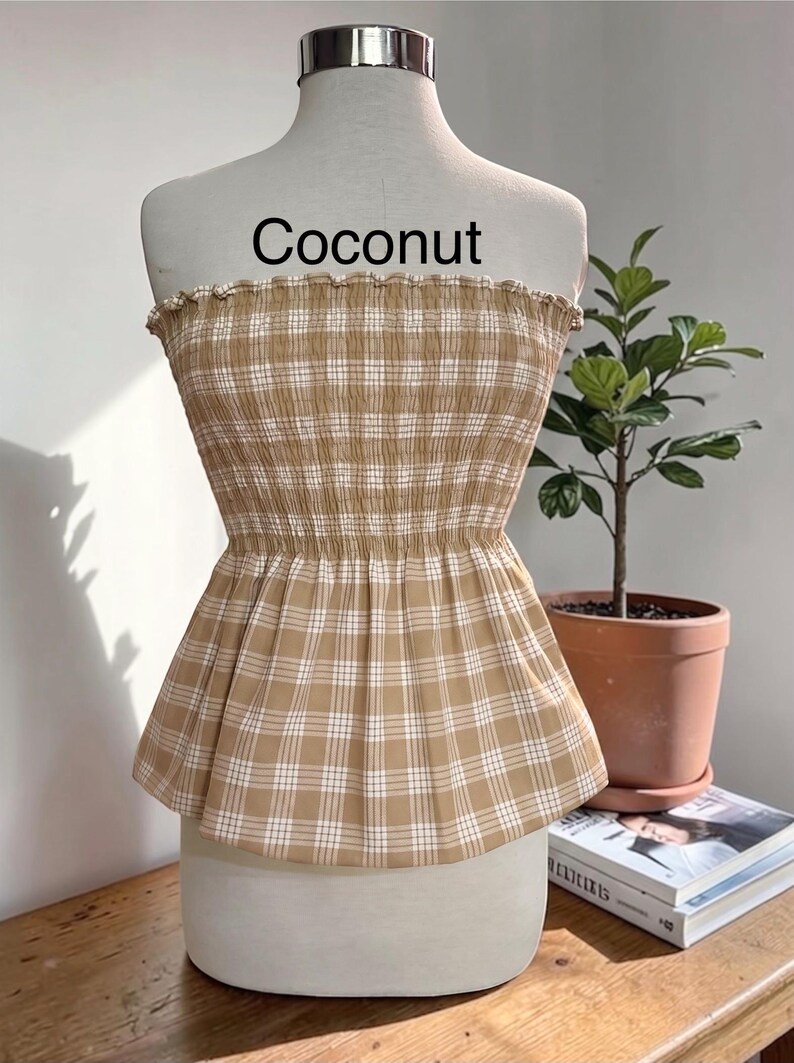 May include: A strapless tube top in a tan and white plaid pattern with a smocked, elasticized bodice and a ruffled top edge. The garment features the word Coconut printed in black text above the neckline on the display mannequin.