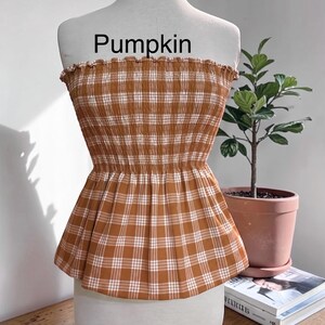 Peut inclure: Un haut bustier sans bretelles avec un motif &agrave; carreaux marron et blanc, dot&eacute; d'un corsage smock&eacute; &eacute;lastique et d'un bord sup&eacute;rieur volant&eacute;. Le mot Pumpkin est imprim&eacute; en noir au-dessus du v&ecirc;tement, pr&eacute;sent&eacute; sur un buste de mannequin blanc.
